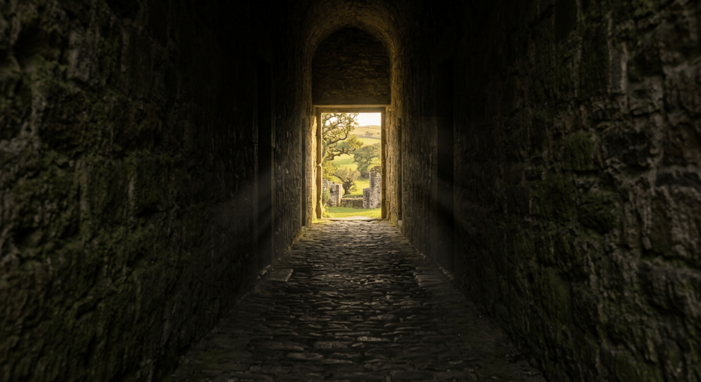 Narrow stone corridor with sunlight at the end revealing a green garden and distant hills